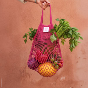 Vor einer pfirsichfarbenen Wand hält eine Hand eine rosa Netz-Tüte, die mit Stangensellerie, einer gelben Melone, roten und grünen Paprikaschoten sowie Tomaten gefüllt ist.