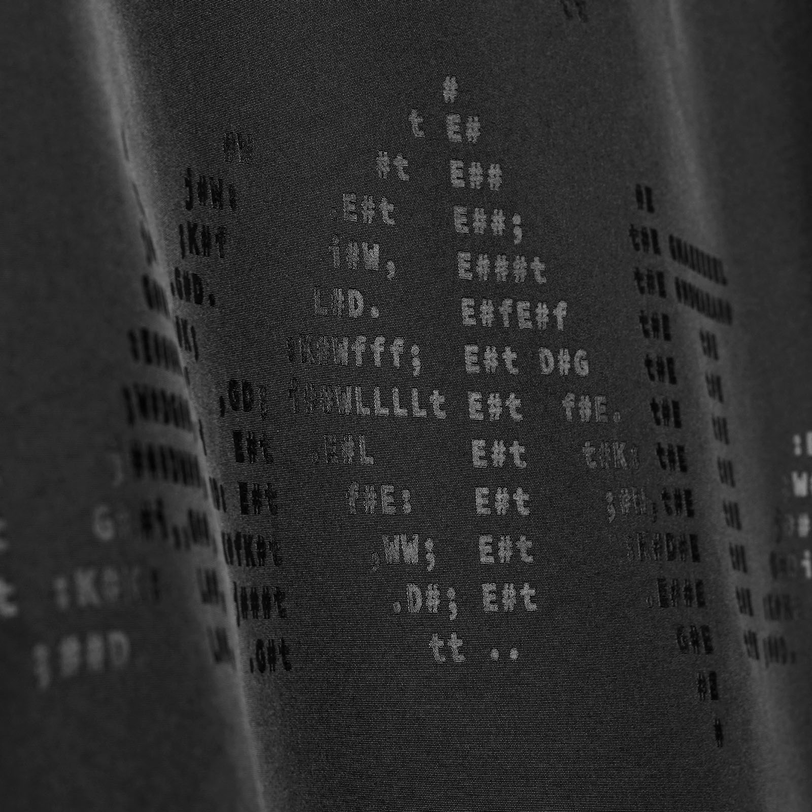 Schwarzer Stoff mit glänzendem, geprägtem Text und Symbolen, die zu einem Muster angeordnet sind. Die Zeichen sind schwer zu entziffern, wodurch auf dem Stoff ein abstrakter, strukturierter Effekt entsteht.