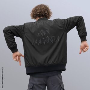 Eine Person mit lockigem Haar trägt eine schwarze Jacke, auf deren Rücken in gepunkteter Schrift die Worte „LOVE MAHANTA“ auf einem schlichten hellgrauen Hintergrund stehen; die Arme sind angewinkelt und zeigen nach unten.