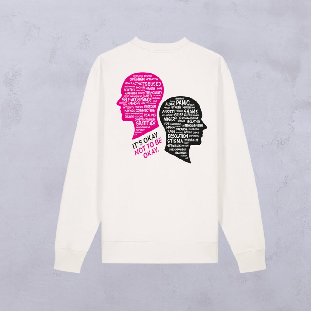 Auf der Rückseite eines weißen Sweatshirts sind zwei sich überlappende Kopf-Silhouetten zu sehen: eine in Rosa mit positiven Worten, die andere in Schwarz mit negativen Worten. Darunter steht der Schriftzug: „ITS OKAY NOT TO BE OKAY“.