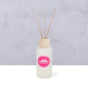 Ein Diffusorflakon aus Milchglas mit einem Holzverschluss und mehreren Holzstäbchen, beschriftet mit „LOVE MAGENTA ROOM FRAGRANCE“, steht auf einer hellen Unterlage vor einem grauen, strukturierten Hintergrund.