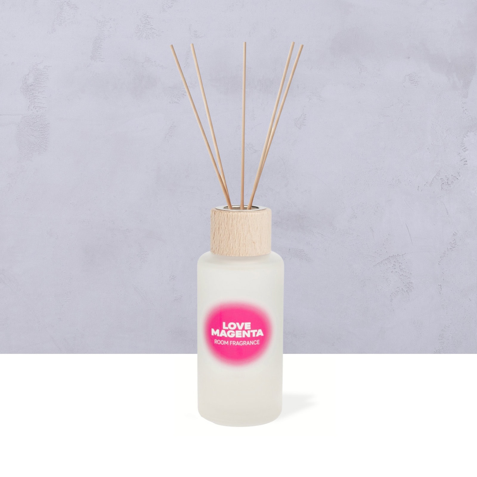 Ein Diffusorflakon aus Milchglas mit einem Holzverschluss und mehreren Holzstäbchen, beschriftet mit „LOVE MAGENTA ROOM FRAGRANCE“, steht auf einer hellen Unterlage vor einem grauen, strukturierten Hintergrund.