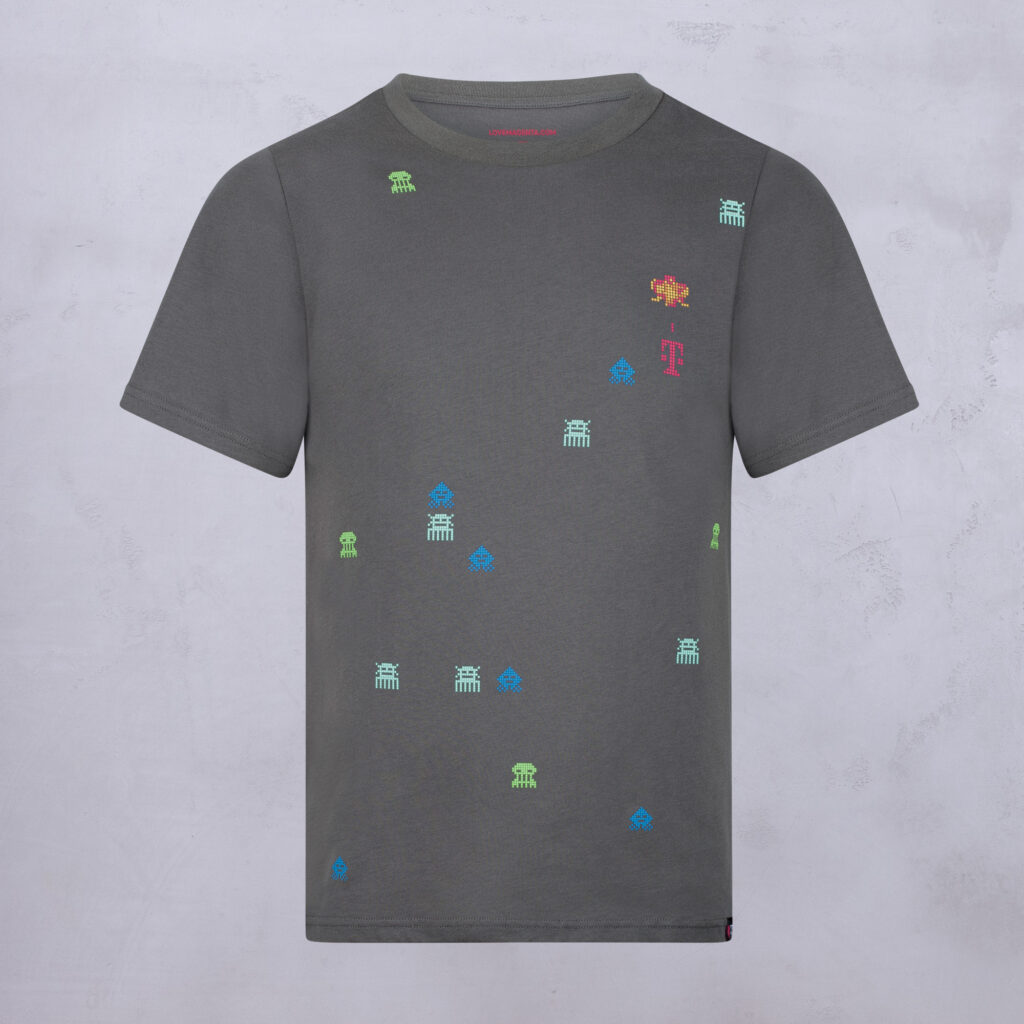 Ein graues T-Shirt, auf dessen Vorderseite bunte, kleine Pixel-Art-Motive mit Außerirdischen und Symbolen verteilt sind. Das Shirt wird vor einem hellgrauen Hintergrund gezeigt.