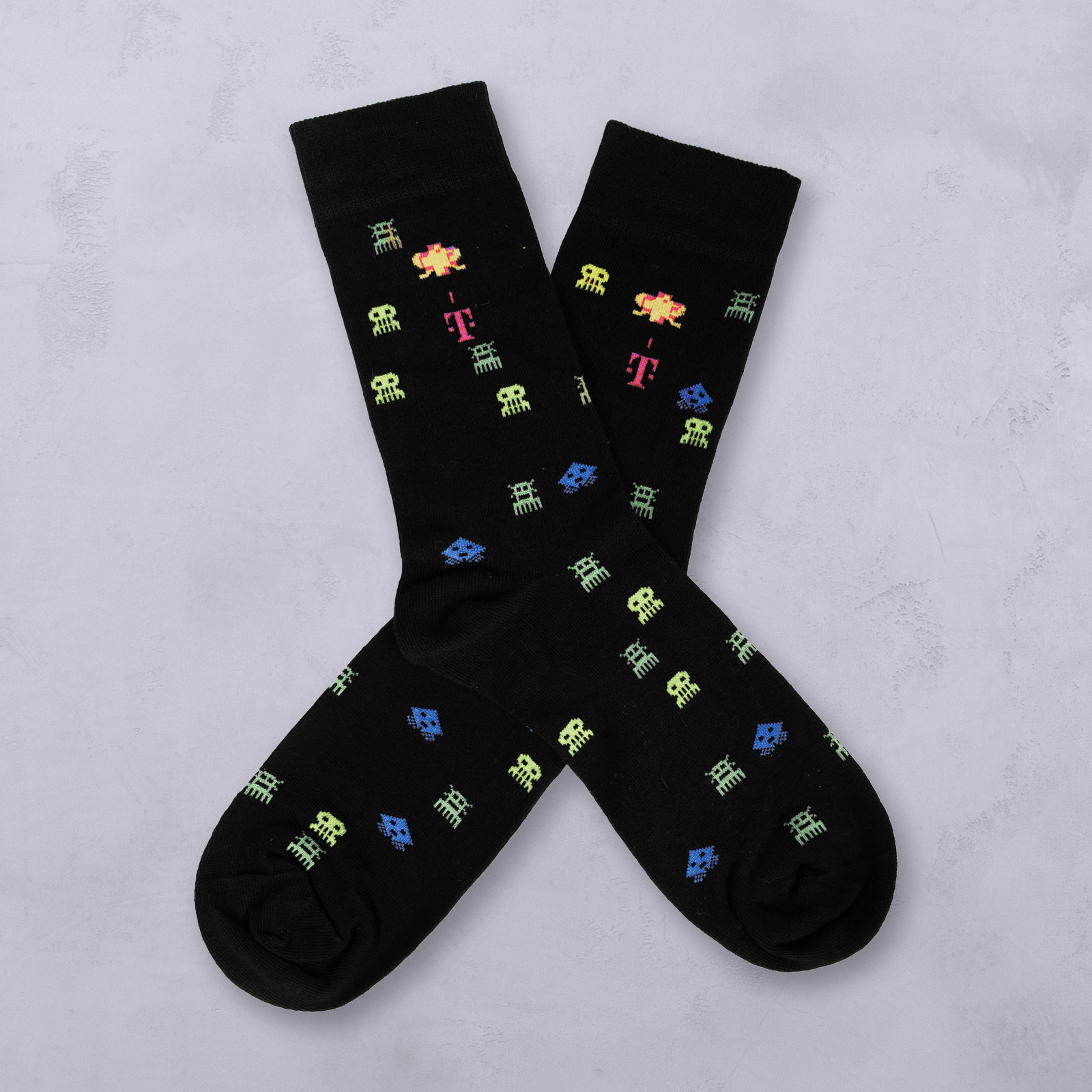 Schwarze Socken mit einem bunten, pixelartigen Muster aus Außerirdischen und Symbolen, darunter grüne, blaue und gelbe Formen sowie einige magentafarbene T-Logos, die sich auf einem hellgrauen, strukturierten Hintergrund überkreuzen.