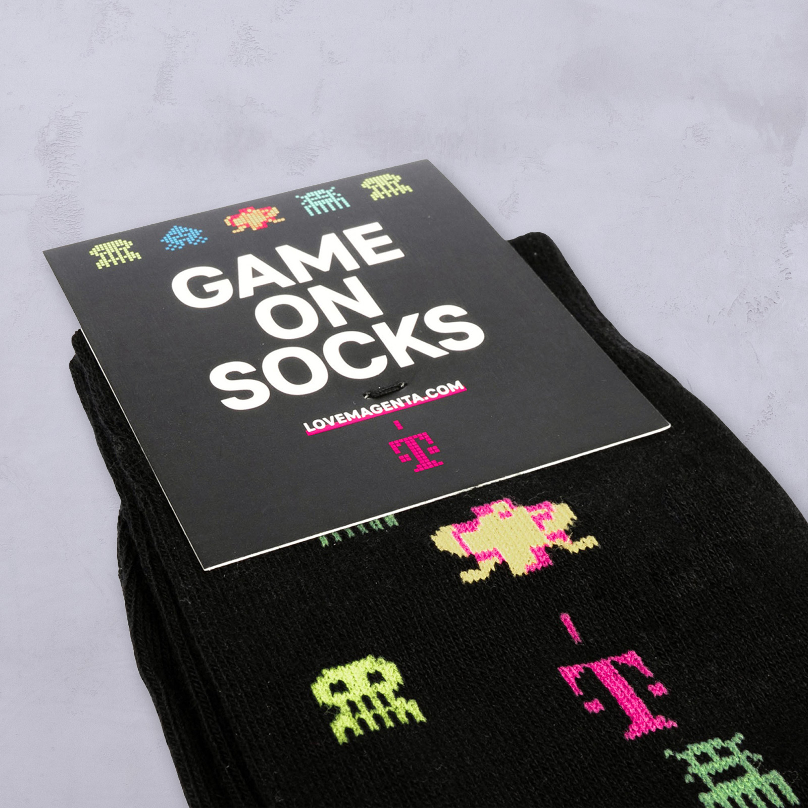 Ein Paar schwarze Socken mit bunten Aliens und Symbolen im Pixel-Art-Stil, verpackt mit einem Etikett, auf dem „GAME ON SOCKS“ und die Website „LOVEMAGENTA.COM“ sowie ein pinkfarbenes T-Mobile-Logo zu sehen sind.