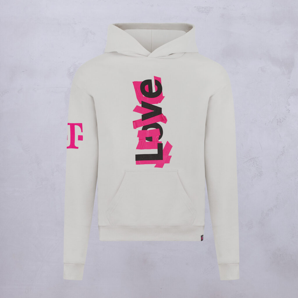 Weißer Hoodie mit dem Wort „Love“ in fetten schwarzen und rosa Buchstaben, die vertikal über die Vorderseite verlaufen; auf dem linken Ärmel ist ein rosa „T“ zu sehen. Der Hintergrund ist eine hellgraue, strukturierte Oberfläche.
