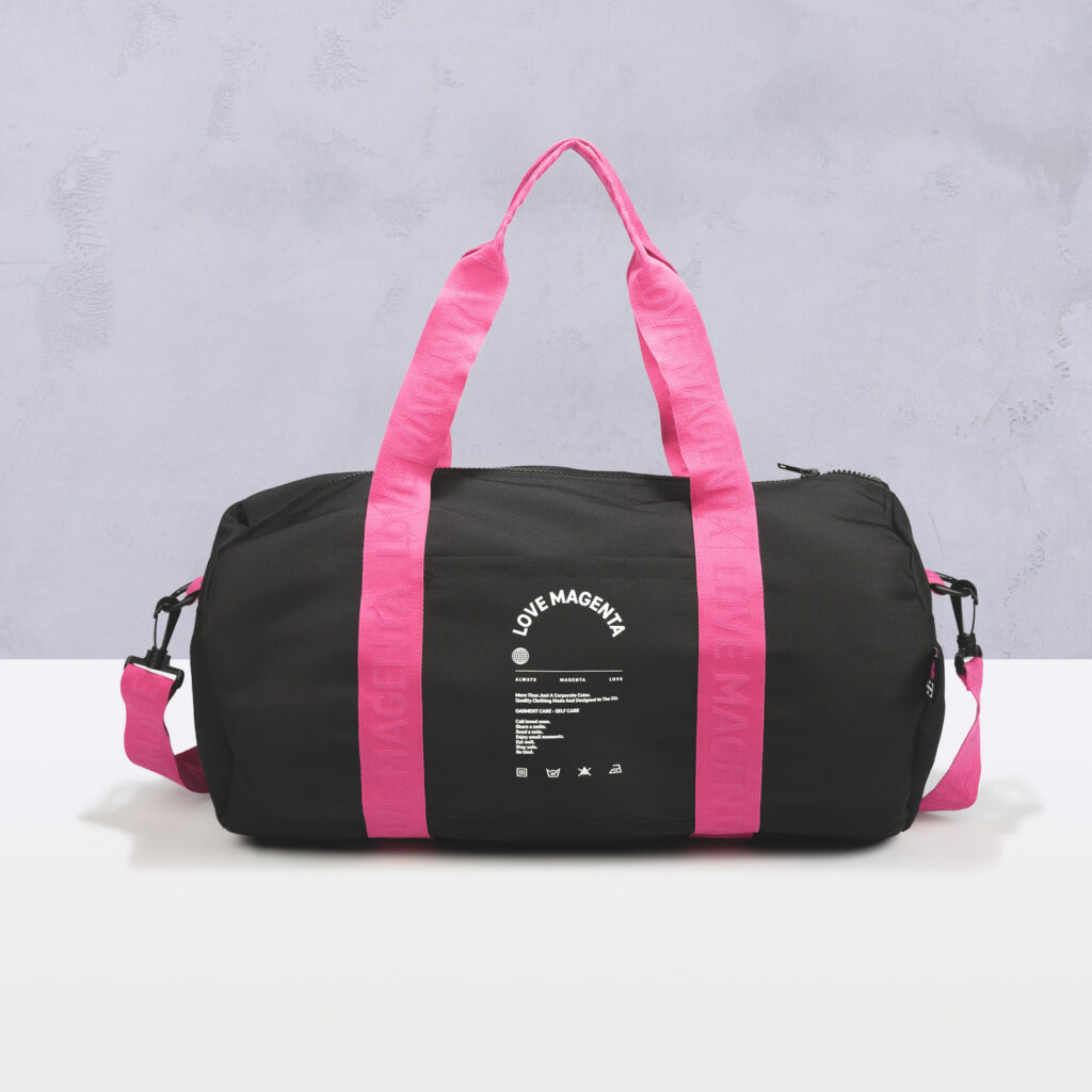Auf einer weißen Fläche vor hellgrauem Hintergrund liegt eine schwarze Reisetasche mit leuchtend pinkfarbenen Griffen und Riemen. An der Seite der Tasche ist in weißer Schrift „LOVE MAGENTA“ aufgedruckt.