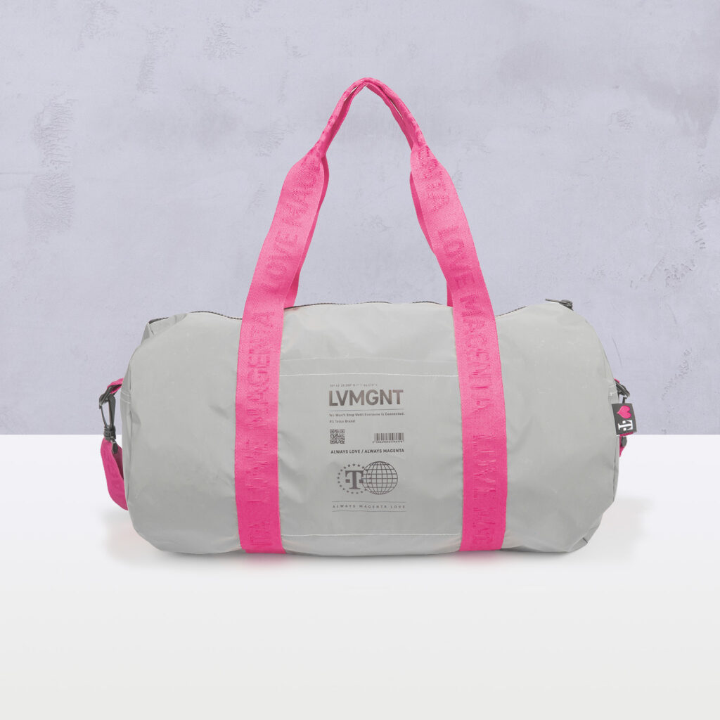 Auf einer weißen Fläche vor einer hellgrauen Wand steht eine hellgraue Reisetasche mit leuchtend pinkfarbenen Griffen und Riemen. Auf der Vorderseite der Tasche sind ein Aufdruck und ein kleiner Barcode zu sehen.