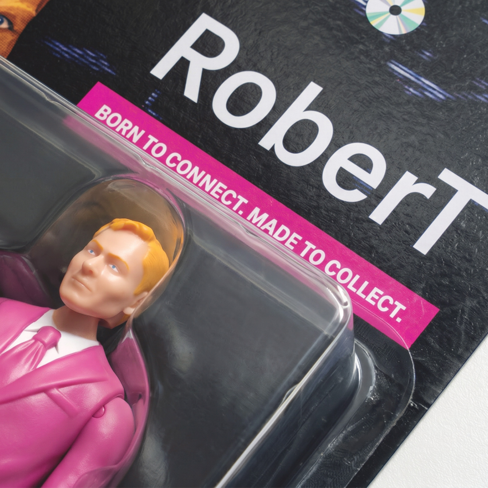 Eine Nahaufnahme einer Sammler-Actionfigur in einem rosa Anzug, verpackt unter dem Markenaufdruck „RoberT“ und „BORN TO CONNECT. MADE TO COLLECT.“ auf schwarzem Hintergrund.