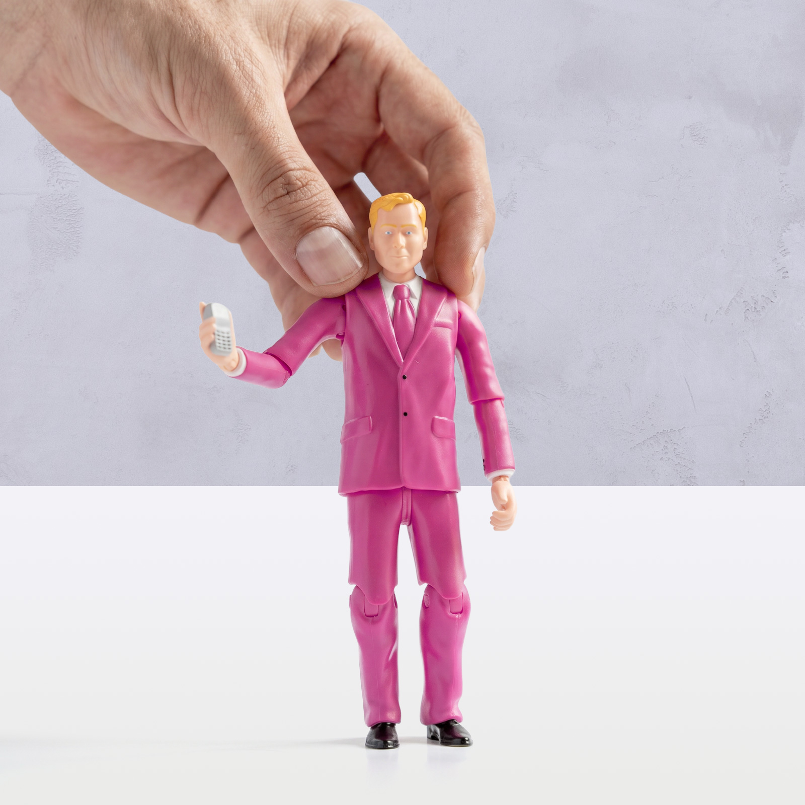 Eine Hand hält eine kleine Actionfigur in einem rosa Anzug, die ein weißes Handy in der Hand hält; das Ganze ist vor einem hellgrauen Hintergrund zu sehen.