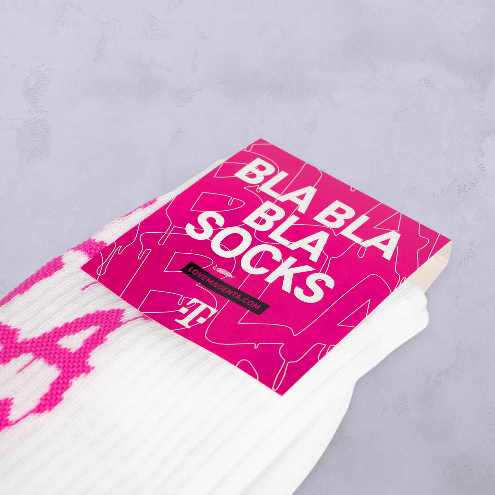 Ein Paar weiße Socken mit einem rosa Etikett, auf dem „BLA BLA BLA SOCKS“ und „LOLAMOSHKA.COM“ auf hellgrauem Hintergrund stehen.