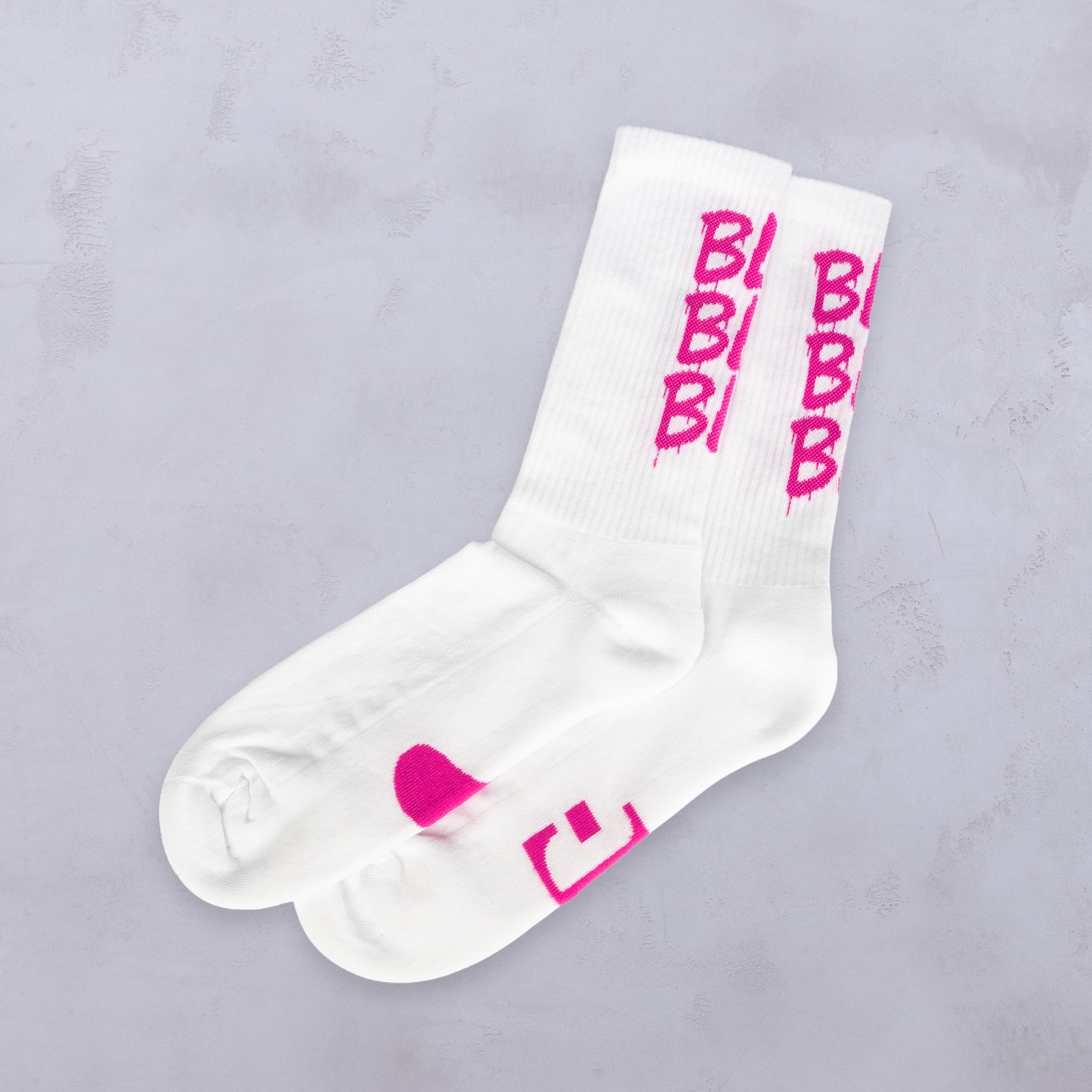 Ein Paar weiße Crew-Socken mit auffälligem pinkfarbenem „BB“-Schriftzug an den Seiten und einem pinkfarbenen Motiv im Zehenbereich, auf einem hellgrauen, strukturierten Untergrund.