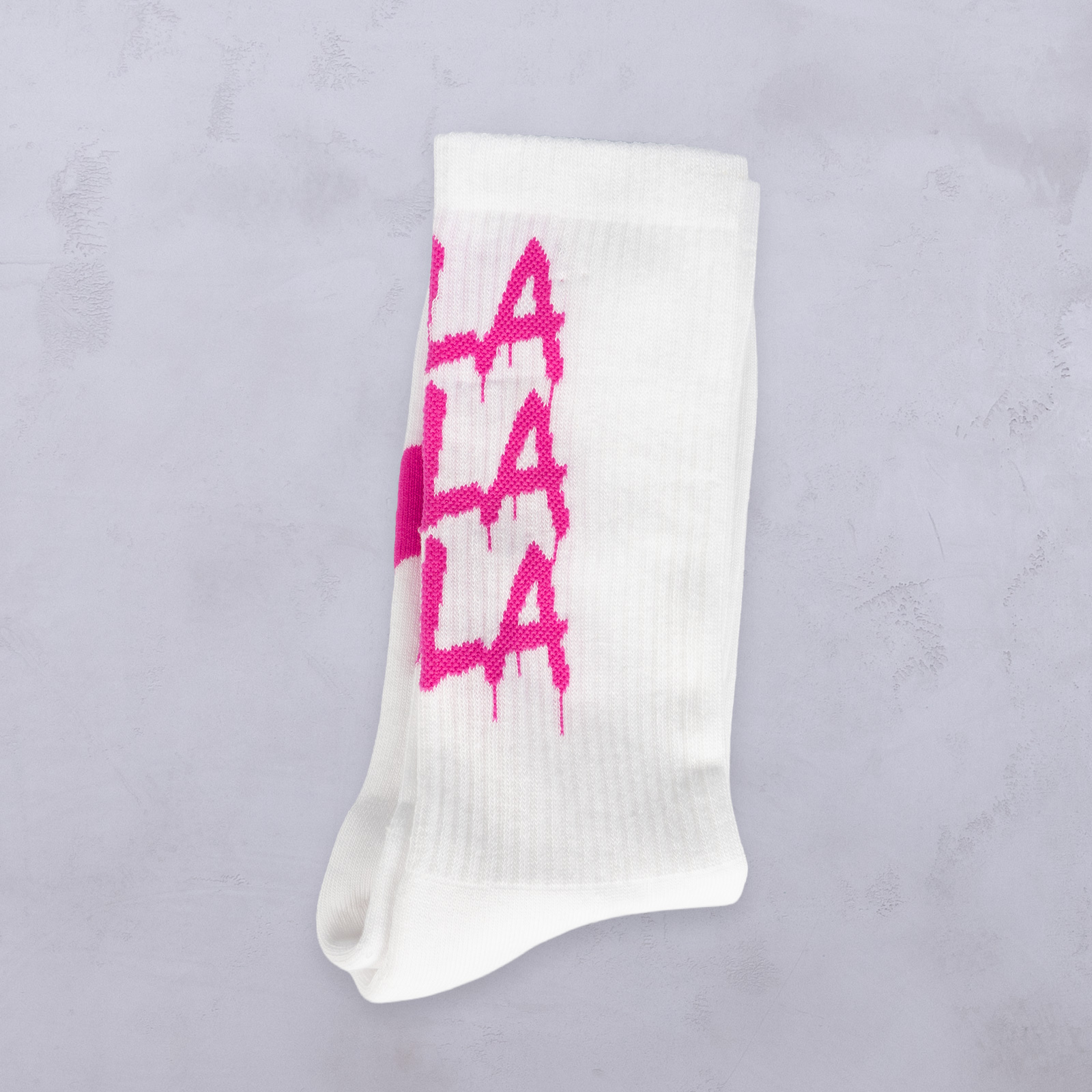 Weiße Socke mit einem auffälligen rosa „LA LA LA LA“-Motiv im Farbtropfen-Stil auf hellgrauem, strukturiertem Untergrund.