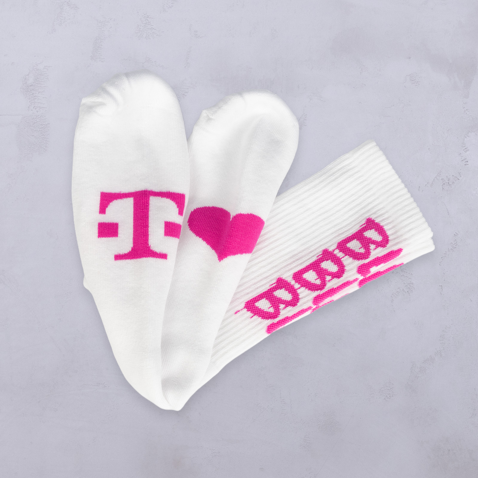 Weiße Socken mit rosa Motiven: Auf einer ist ein „T“ zu sehen, auf einer anderen ein Herz und auf der dritten sind Brezelformen abgebildet. Alle sind auf einem hellgrauen Hintergrund angeordnet.