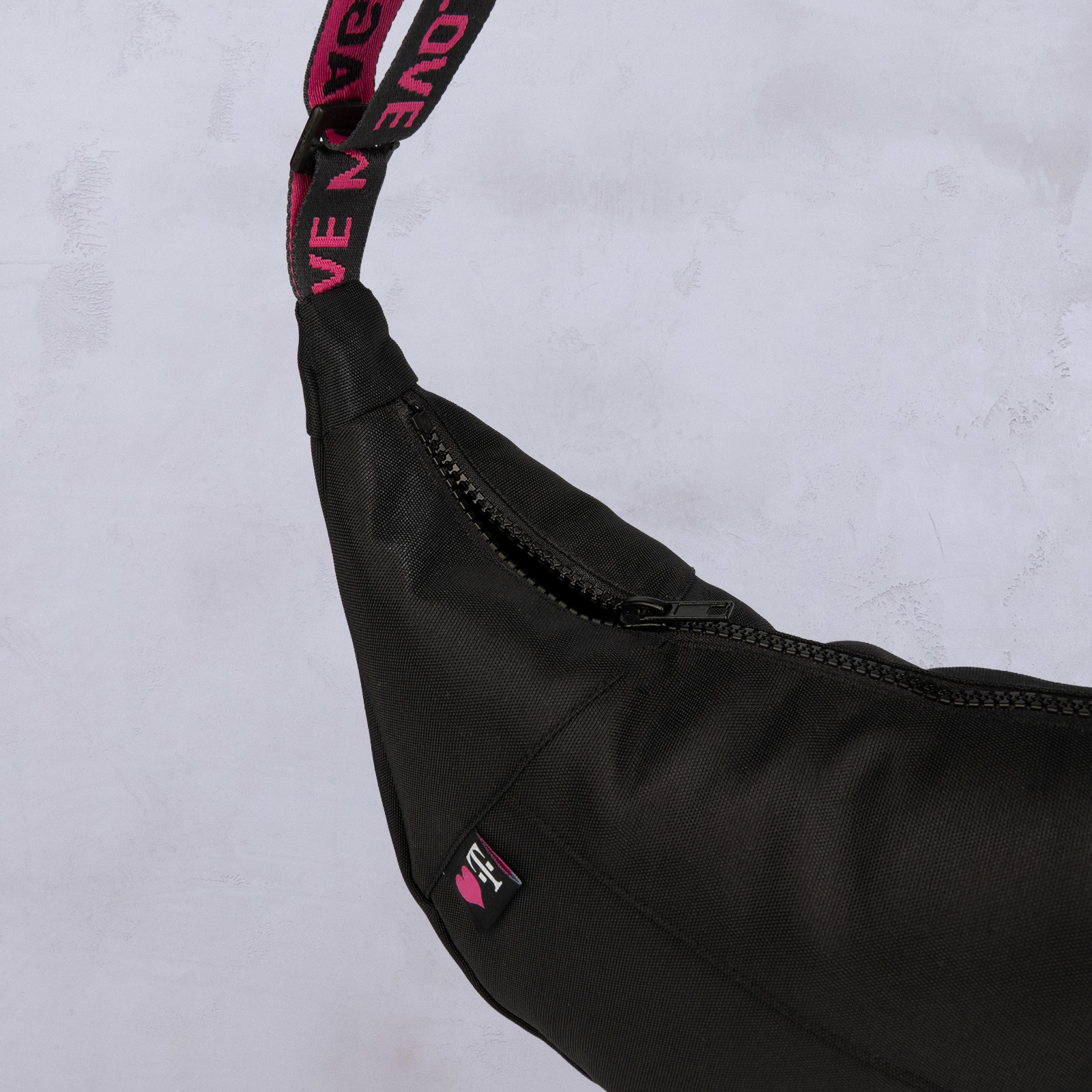 Schwarze Umhängetasche mit einem rosa-schwarzen Riemen, auf dem das Wort „LOVE“ zu sehen ist. Die Tasche ist leicht geöffnet, sodass der Reißverschluss sichtbar ist, und befindet sich vor einem hellgrauen Hintergrund.