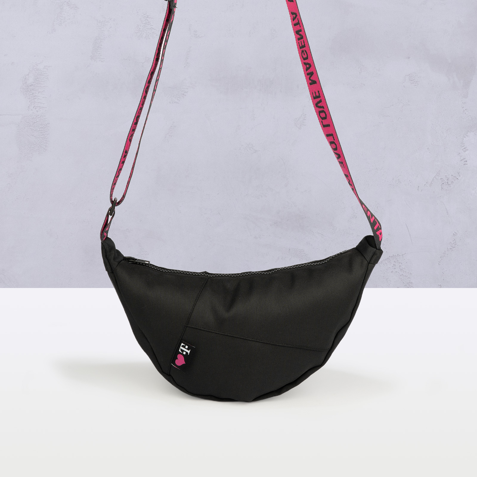 Eine schwarze, halbmondförmige Umhängetasche mit einem rosa Riemen und einem kleinen Aufnäher auf der Vorderseite, abgebildet vor einem hellen Hintergrund.