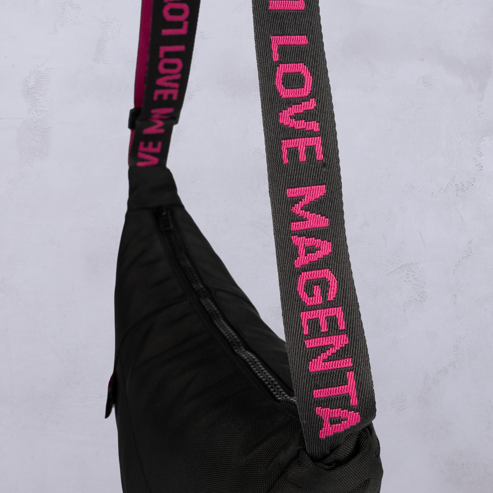 Eine schwarze Tasche mit einem schräg verlaufenden Reißverschluss. Der Trageriemen ist schwarz mit rosa Kanten und einer rosa Stickerei mit der Aufschrift „LOVE MAGENTA“ auf hellgrauem Hintergrund.
