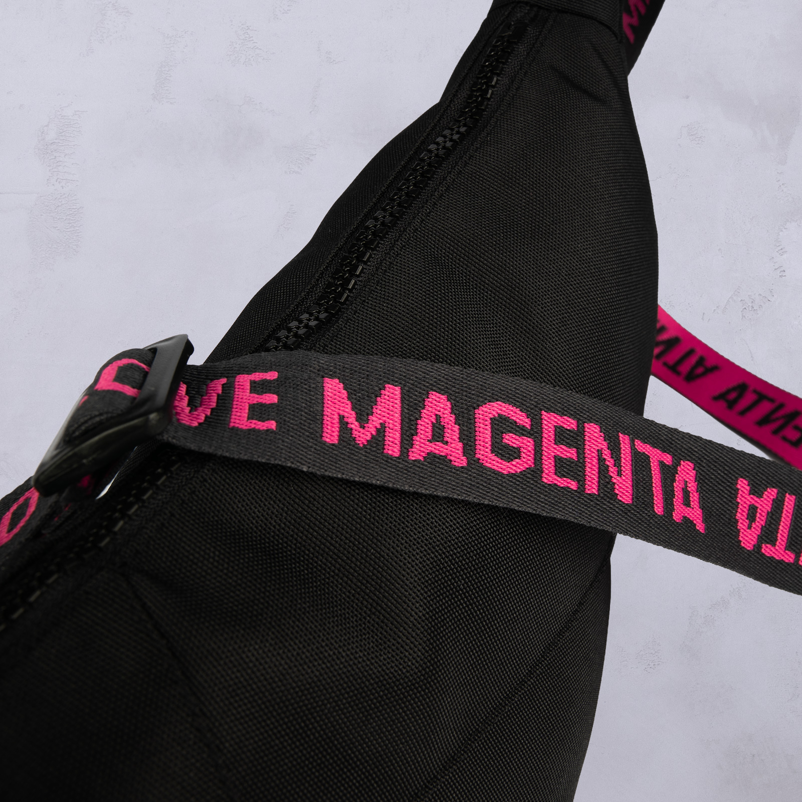 Eine Nahaufnahme einer schwarzen Tasche mit einem schwarzen Trageriemen, auf dem in kräftigem Pink der Schriftzug „MAGENTA“ in Großbuchstaben zu lesen ist. Der Hintergrund ist eine hellgraue, strukturierte Oberfläche.