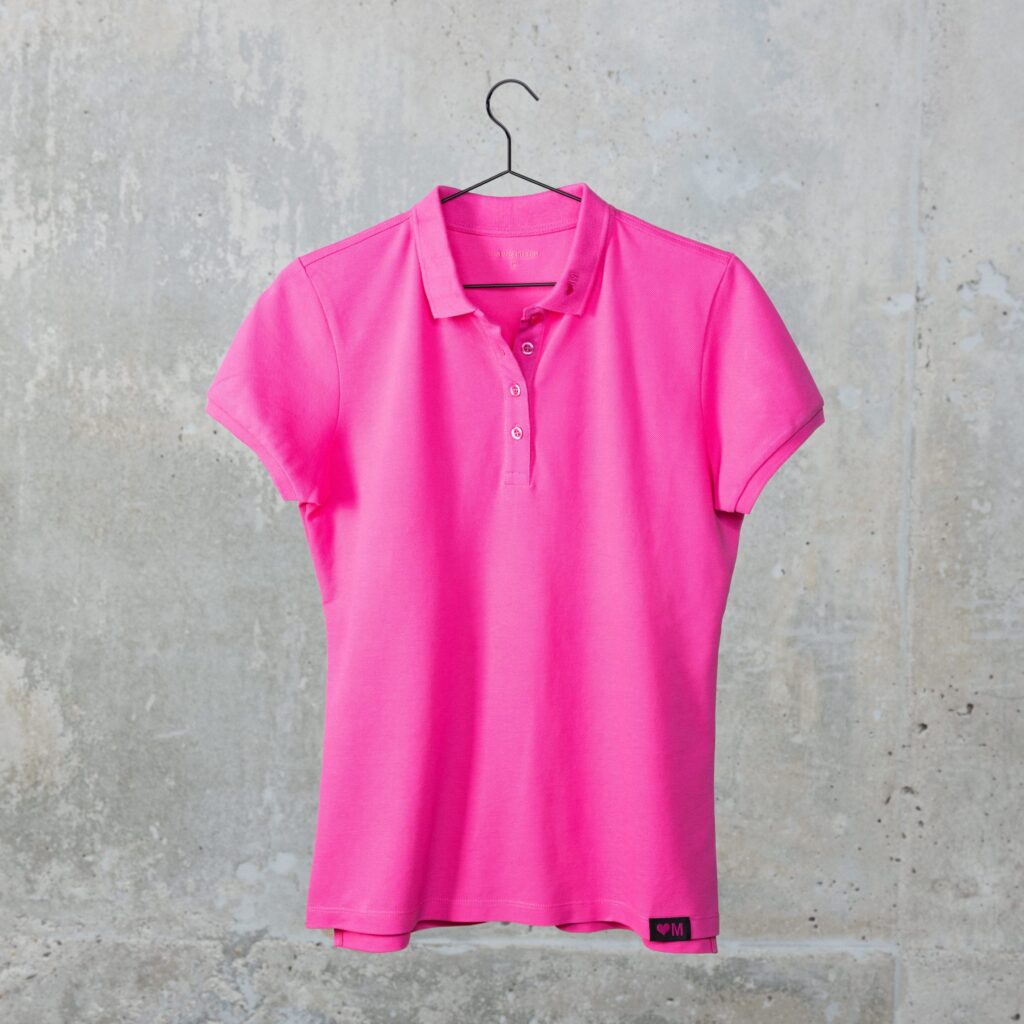 Ein leuchtend pinkfarbenes Kurzarm-Poloshirt hängt an einem schwarzen Kleiderbügel vor dem Hintergrund einer schlichten Betonwand.