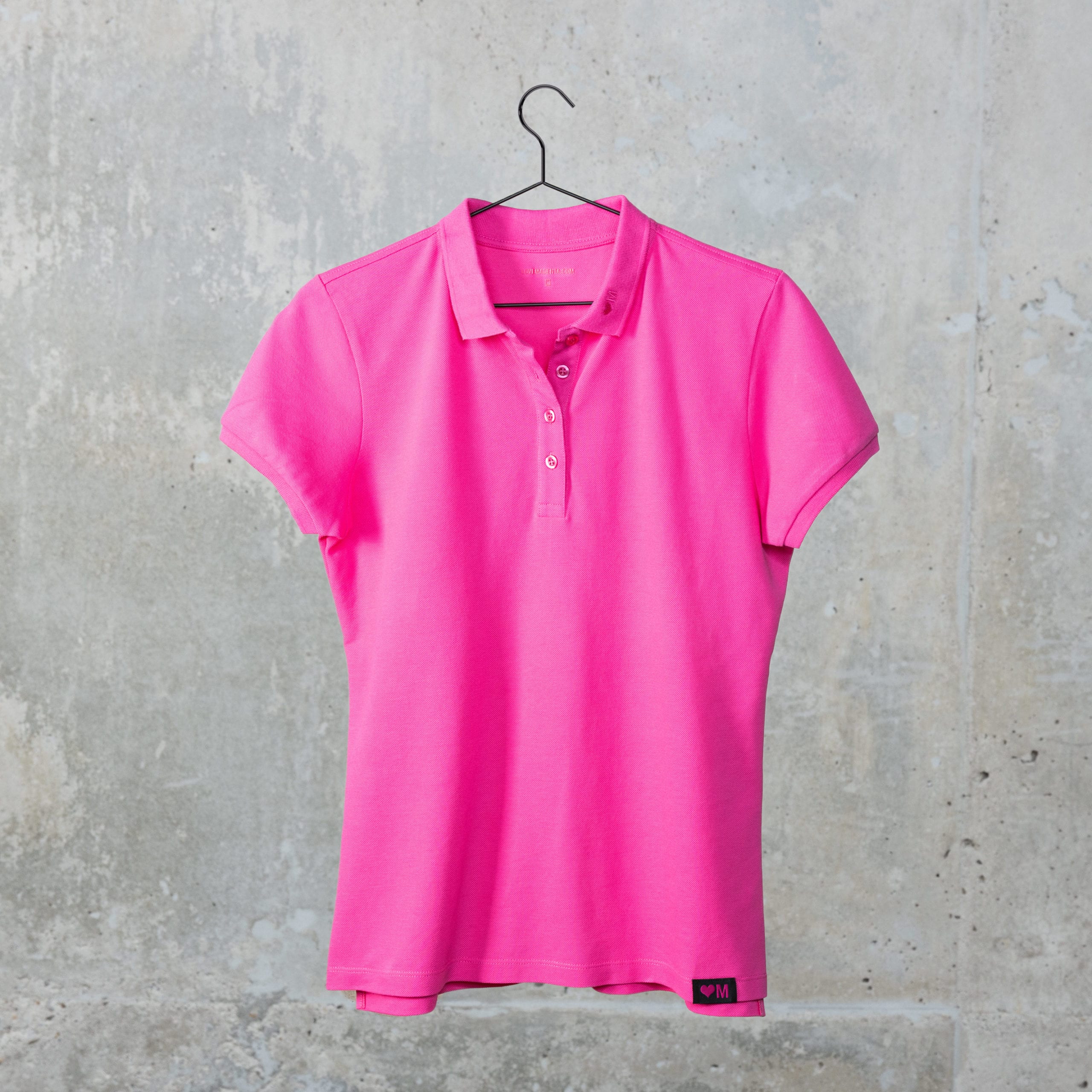 Ein leuchtend pinkfarbenes Kurzarm-Poloshirt hängt an einem schwarzen Kleiderbügel vor dem Hintergrund einer schlichten Betonwand.
