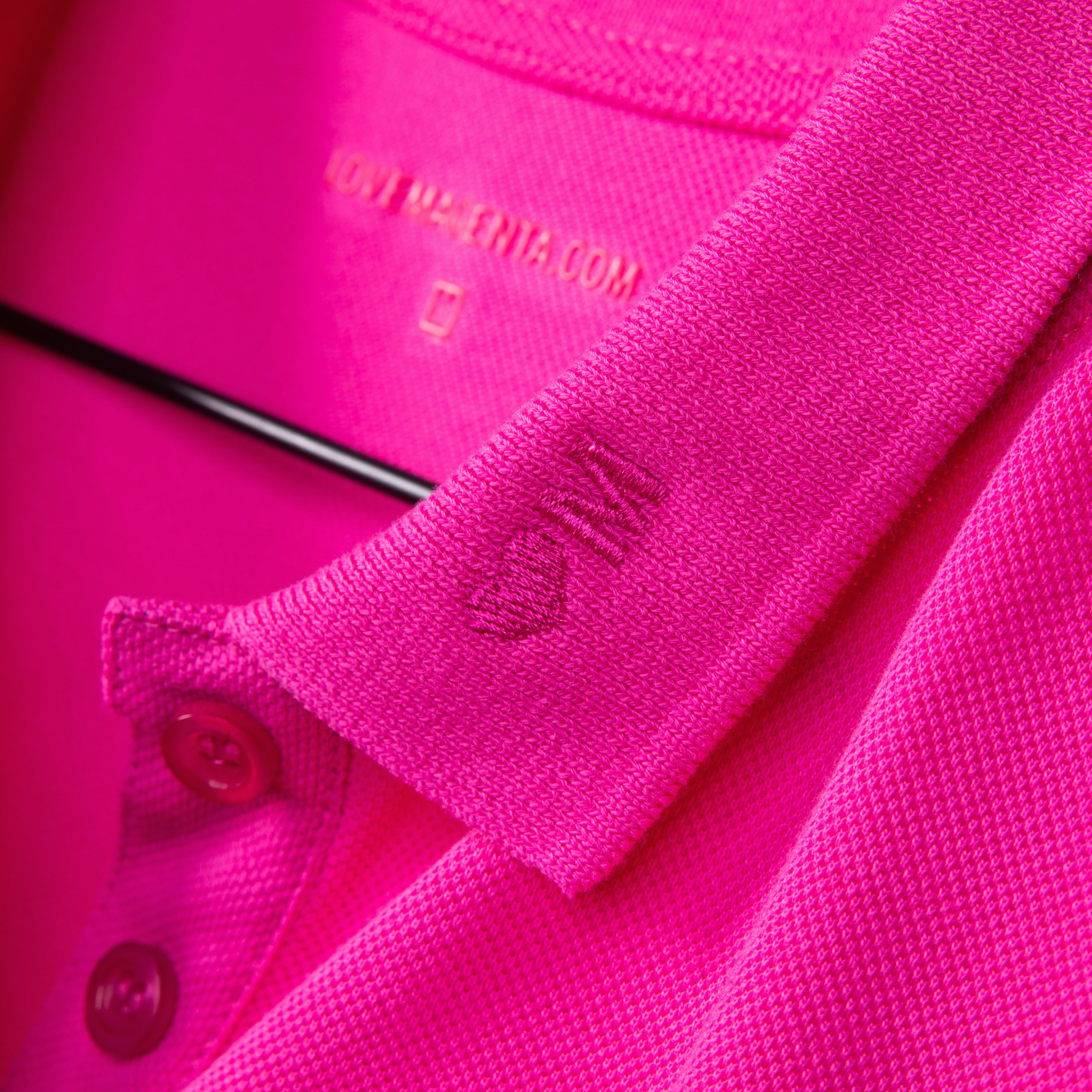Nahaufnahme eines leuchtend rosa Hemds mit Kragen auf einem Kleiderbügel, die die Struktur des Stoffes, die Knöpfe sowie ein kleines gesticktes Herz und ein „M“ am Kragen zeigt.