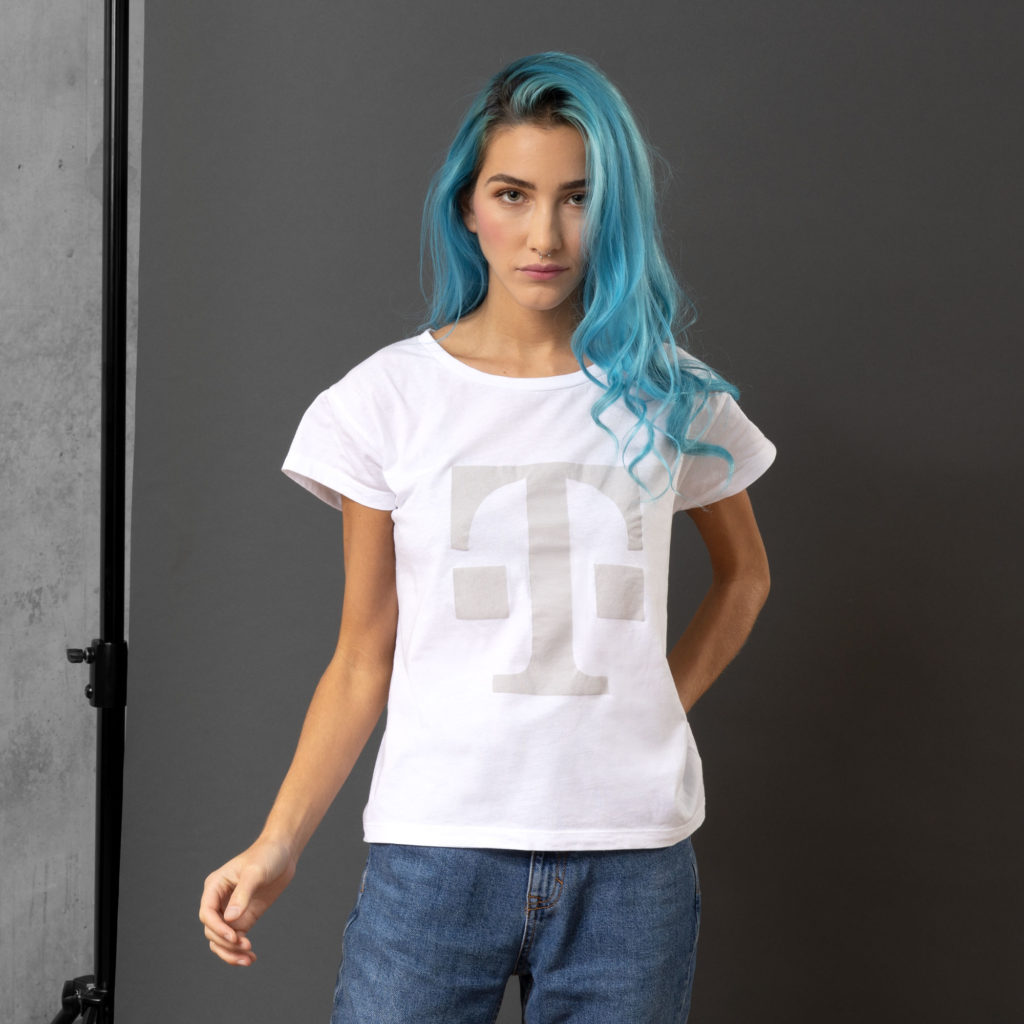 Eine Person mit langen blauen Haaren trägt ein weißes T-Shirt mit einem großen T-Logo und eine blaue Jeans und steht vor einem grauen Hintergrund, wobei sie einen Arm leicht angehoben hat.