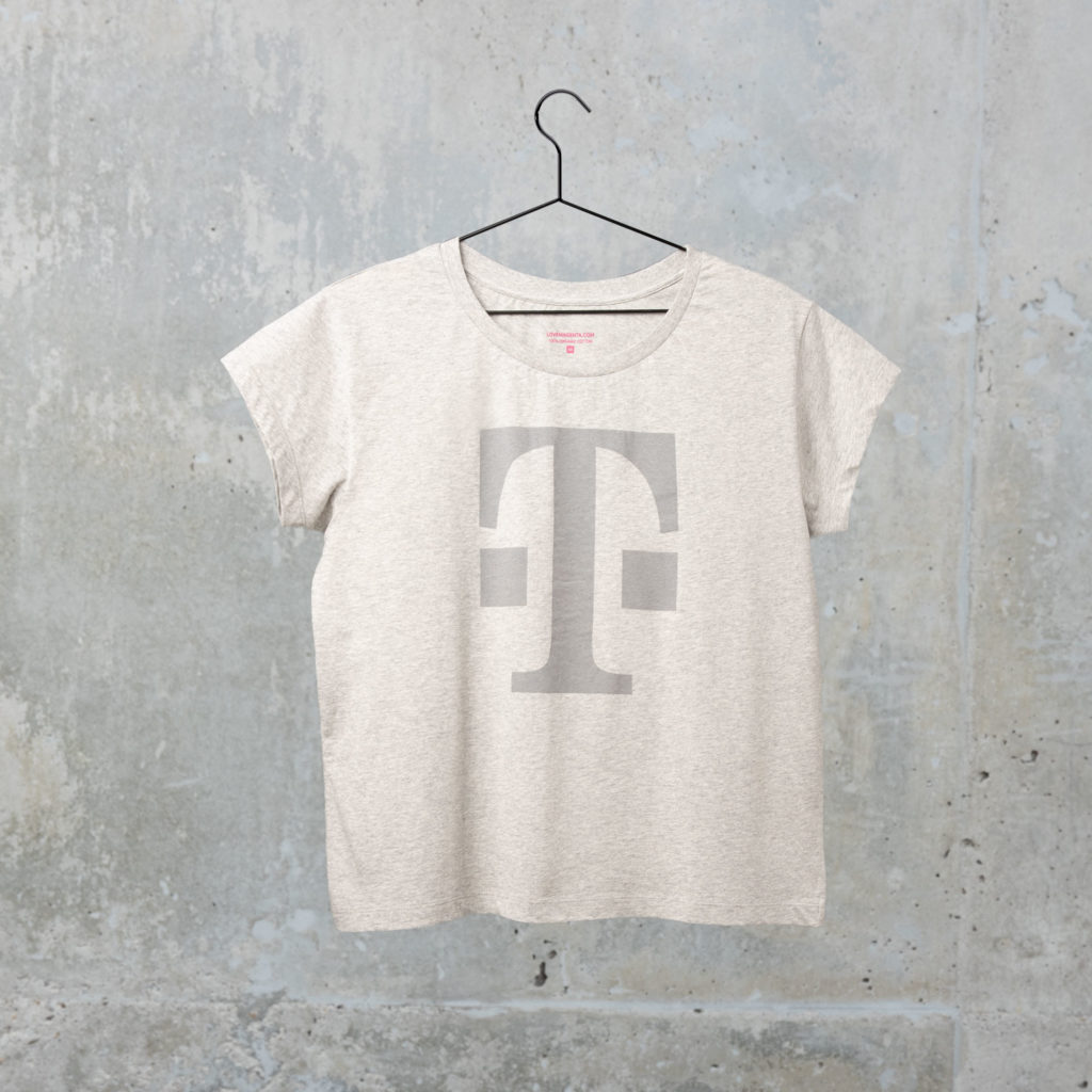 Ein graues T-Shirt mit einem großen, hellgrauen T-Logo auf der Vorderseite, das vor dem Hintergrund einer strukturierten Betonwand an einem schwarzen Kleiderbügel hängt.