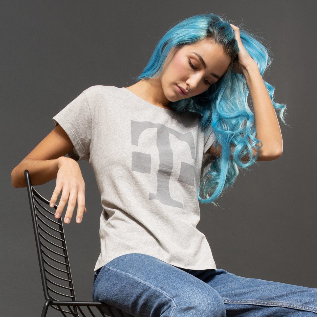 Vor einem dunkelgrauen Hintergrund sitzt eine Person mit langen blauen Haaren auf einem modernen schwarzen Stuhl; sie trägt ein hellgraues T-Shirt mit einem großen Buchstabenmotiv und eine blaue Jeans und berührt mit einer Hand ihr Haar.