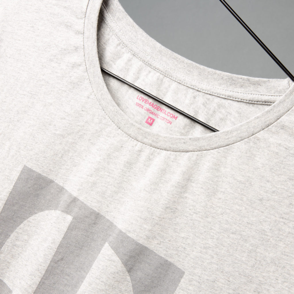 Nahaufnahme eines hellgrauen T-Shirts auf einem Kleiderbügel, auf der der Ausschnitt und ein rosa Etikett mit der Aufschrift „LOVE BACK ENZY.COM 100 % ORGANIC COTTON M“ zu sehen sind.