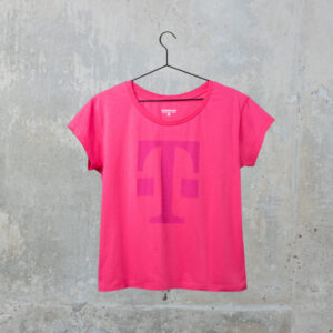 Ein rosa T-Shirt mit einem großen magentafarbenen „T“ auf der Vorderseite hängt an einem schwarzen Kleiderbügel vor einer grauen Betonwand.