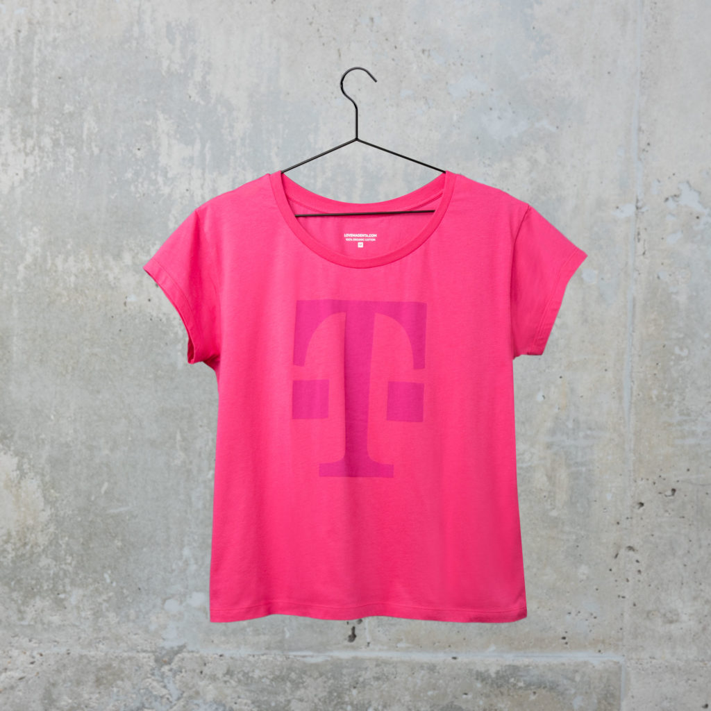 Ein rosa T-Shirt mit einem großen magentafarbenen „T“ auf der Vorderseite hängt an einem schwarzen Kleiderbügel vor einer grauen Betonwand.
