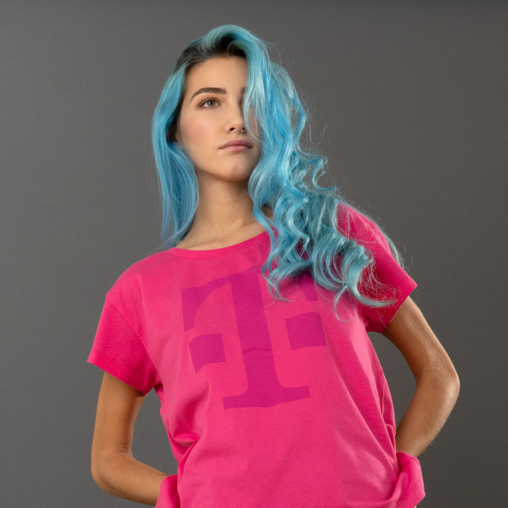 Eine Person mit langen blauen Haaren, die ein leuchtend pinkes T-Shirt mit einem großen violetten „T“ trägt, steht vor einem schlichten grauen Hintergrund und blickt leicht von der Kamera weg.