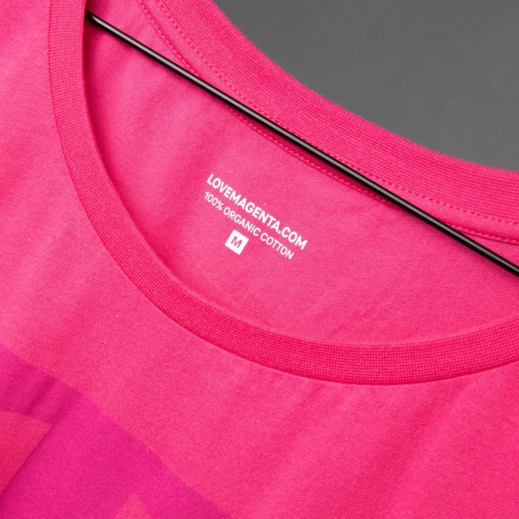Nahaufnahme eines rosa T-Shirts auf einem Kleiderbügel, auf der der Ausschnitt und ein aufgedrucktes Etikett mit der Aufschrift „LOVEMAGENTA.COM, 100 % BIO-BAUMWOLLE, M“ in weißer Schrift zu sehen sind.
