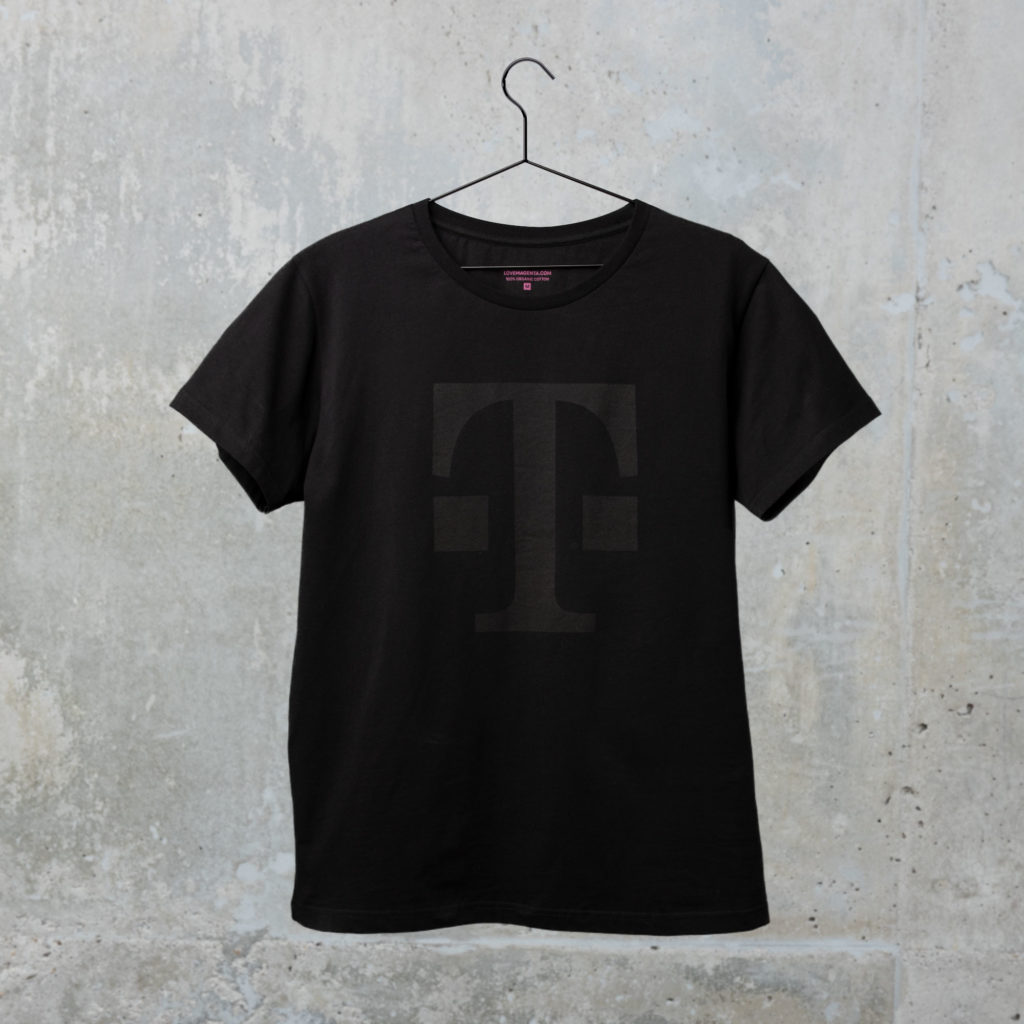 Ein schwarzes T-Shirt mit einem großen, dezenten, dunklen „T“-Logo auf der Vorderseite, das an einem schwarzen Kleiderbügel vor einer hellgrauen Betonwand hängt.