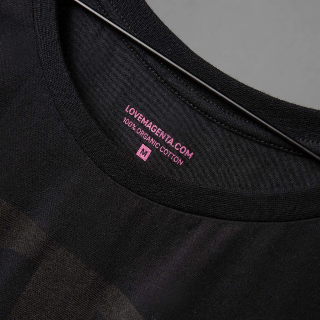 Nahaufnahme eines schwarzen T-Shirts auf einem Kleiderbügel, auf dem im Kragenbereich ein rosa Etikett zu sehen ist, auf dem „LOVEMAGENTA.COM, 100 % BIO-BAUMWOLLE, M“ steht.