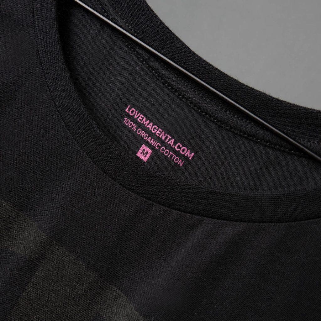 Nahaufnahme eines schwarzen T-Shirts auf einem Kleiderbügel, auf dem im Kragenbereich ein rosa Etikett mit der Aufschrift „LOVEMAGENTA.COM 100 % ORGANIC COTTON M“ zu sehen ist.