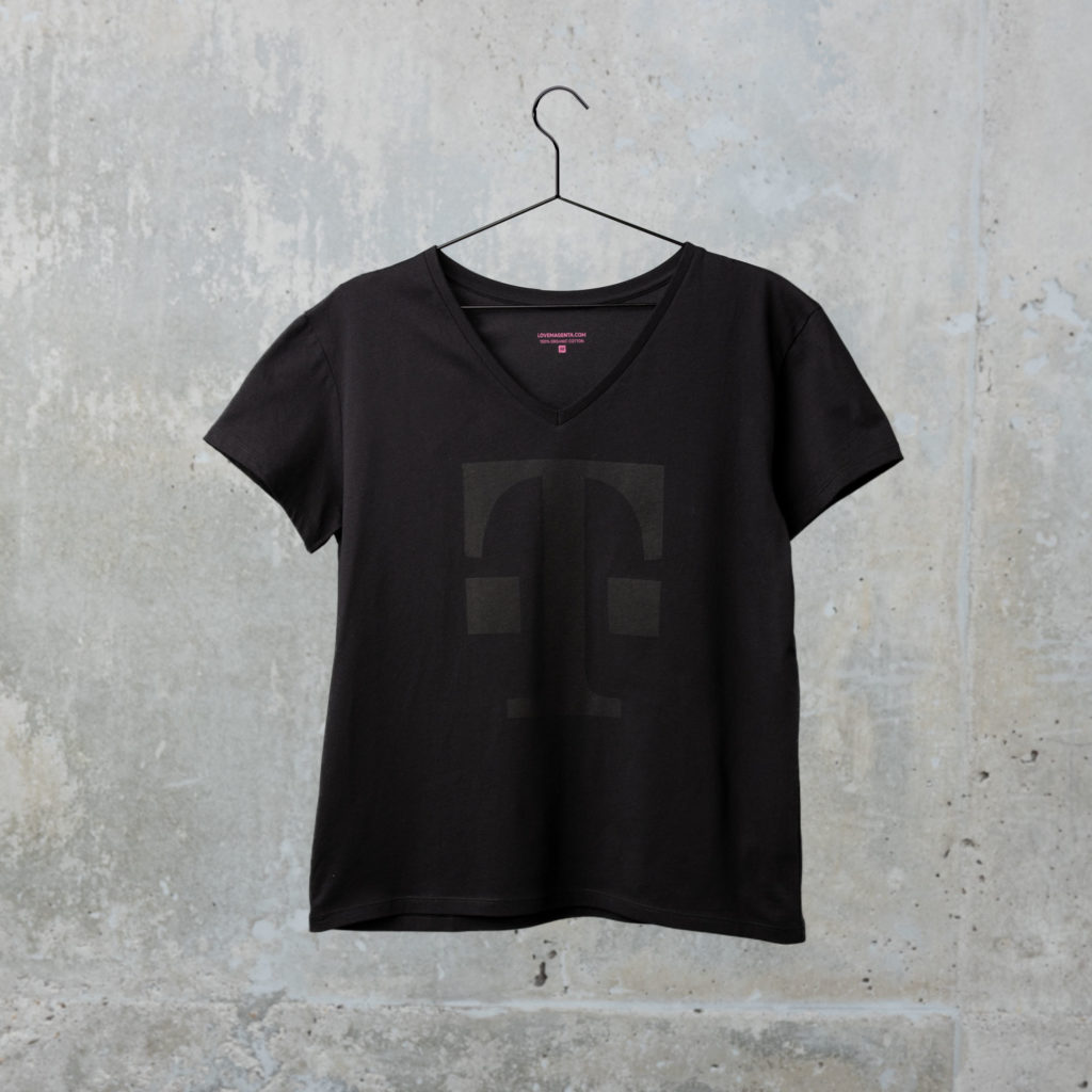Ein schwarzes T-Shirt mit V-Ausschnitt und einem großen, dunklen, stilisierten T-Logo auf der Vorderseite hängt an einem schwarzen Kleiderbügel vor dem Hintergrund einer hellgrauen Betonwand.