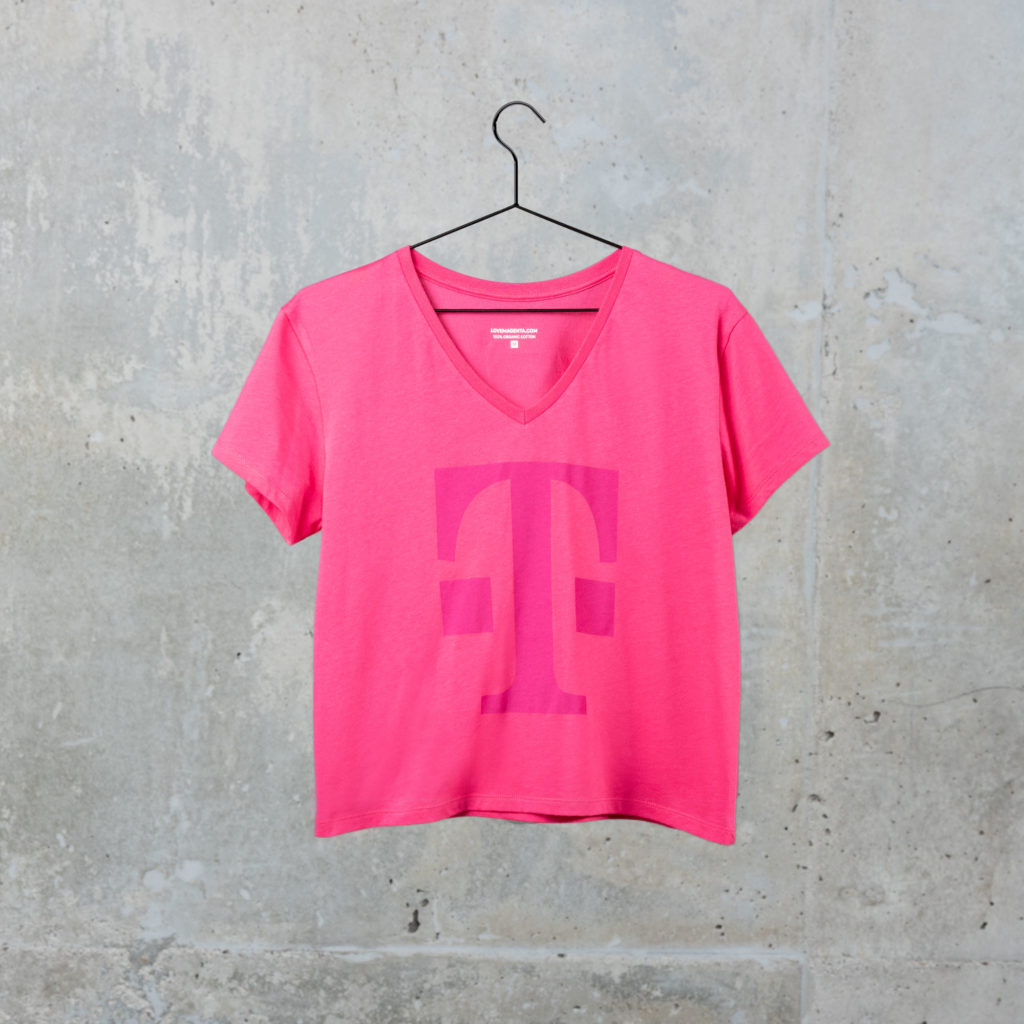 Ein rosa T-Shirt mit einem großen, magentafarbenen „T“-Logo auf der Vorderseite hängt an einem schwarzen Kleiderbügel vor einer grauen Betonwand.