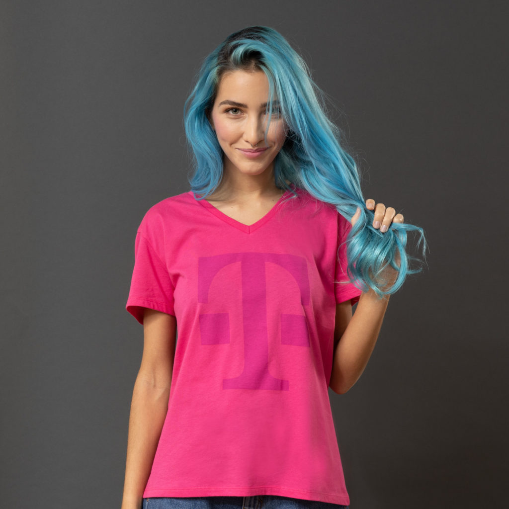 Eine Person mit langen blauen Haaren, die ein rosa T-Shirt mit einem großen lila „T“ darauf trägt, steht vor einem grauen Hintergrund, lächelt und hält eine Haarsträhne in der Hand.