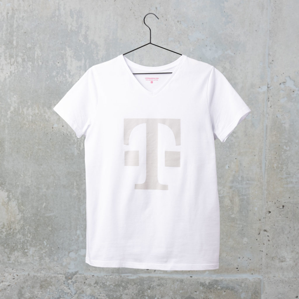 Ein weißes T-Shirt mit einem großen, hellgrauen T-Logo auf der Vorderseite hängt an einem schwarzen Kleiderbügel vor dem Hintergrund einer strukturierten Betonwand.