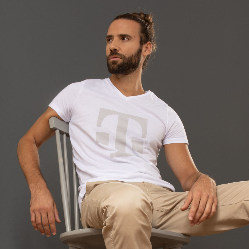 Vor einem schlichten grauen Hintergrund sitzt ein Mann mit Bart und nach hinten gebundenen Haaren seitlich auf einem grauen Stuhl; er trägt ein weißes T-Shirt mit einem großen, hellen Logo und eine beige Hose.