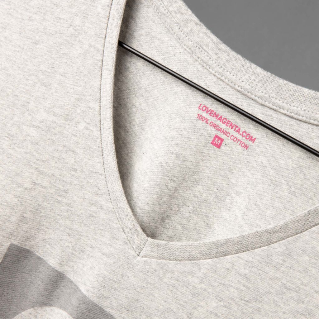 Eine Nahaufnahme eines hellgrauen T-Shirts mit V-Ausschnitt auf einem Kleiderbügel, auf der das Innenetikett mit rosa Schrift zu sehen ist, auf dem „LOVEMAGENTA.COM 100 % BIO-BAUMWOLLE M“ steht.