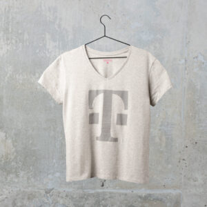 Ein hellgraues T-Shirt mit V-Ausschnitt und einem großen, auffälligen „T“-Motiv auf der Vorderseite hängt an einem schwarzen Kleiderbügel vor einer strukturierten grauen Betonwand.