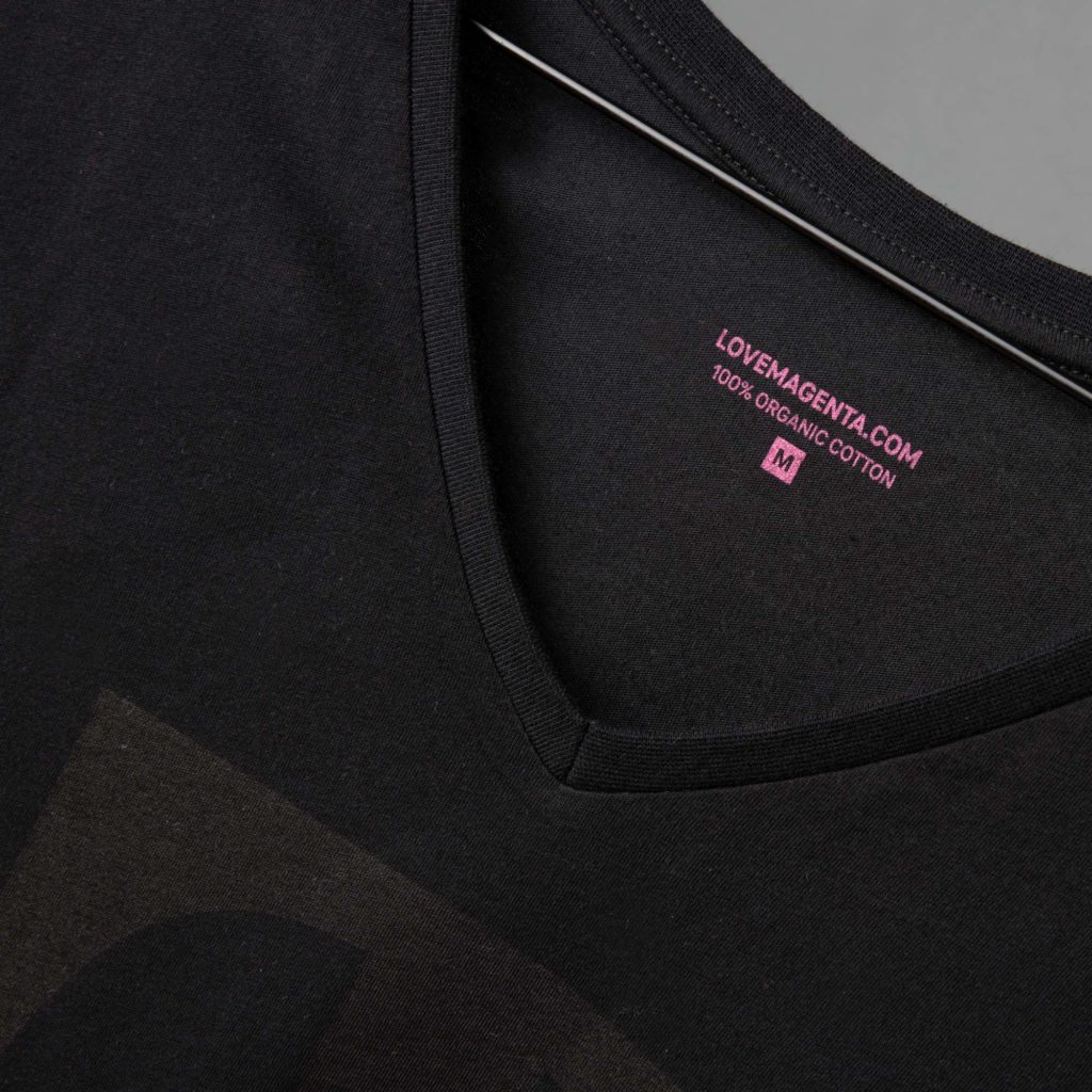 Nahaufnahme eines schwarzen T-Shirts mit V-Ausschnitt auf einem Kleiderbügel, auf dem im Kragenbereich ein rosa Etikett mit der Aufschrift „LOVEMAGENTA.COM 100 % BIO-BAUMWOLLE M“ zu sehen ist.
