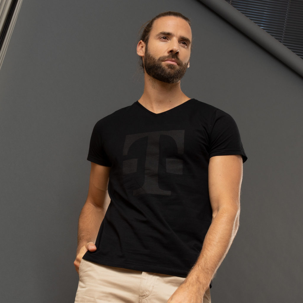 Ein Mann mit Bart und nach hinten gebundenen Haaren trägt ein schwarzes T-Shirt mit dezentem Aufdruck und eine beige Hose; er steht vor einem grauen Hintergrund und hat eine Hand in der Tasche.