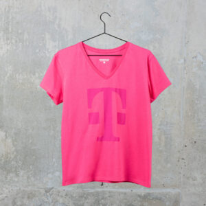 Ein rosa T-Shirt mit V-Ausschnitt und einem großen, stilisierten magentafarbenen „T“-Logo auf der Vorderseite hängt an einem schwarzen Kleiderbügel vor dem Hintergrund einer Betonwand.