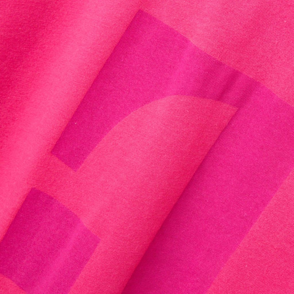 Eine Nahaufnahme von rosa Stoff mit einem dunkleren rosa Aufdruck, der teilweise geometrische Formen bildet – möglicherweise einen Buchstaben oder ein Symbol –, wobei Stofffalten zu erkennen sind.