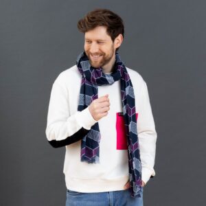 Ein Mann mit braunen Haaren und Bart, der ein weißes Sweatshirt mit einem rosa Quadrat, blaue Jeans und einen gemusterten marineblauen Schal trägt, lächelt und blickt vor einem grauen Hintergrund nach links.