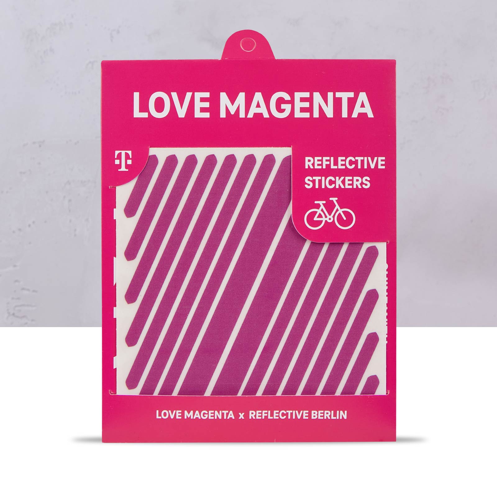 Ein magentafarbenes Paket mit der Aufschrift „LOVE MAGENTA“ enthält reflektierende Aufkleber mit diagonalen magentafarbenen Streifen, die für Fahrräder bestimmt sind. Auf der Verpackung sind das Telekom-Logo und ein Hinweis auf die Zusammenarbeit mit Reflective Berlin zu sehen.