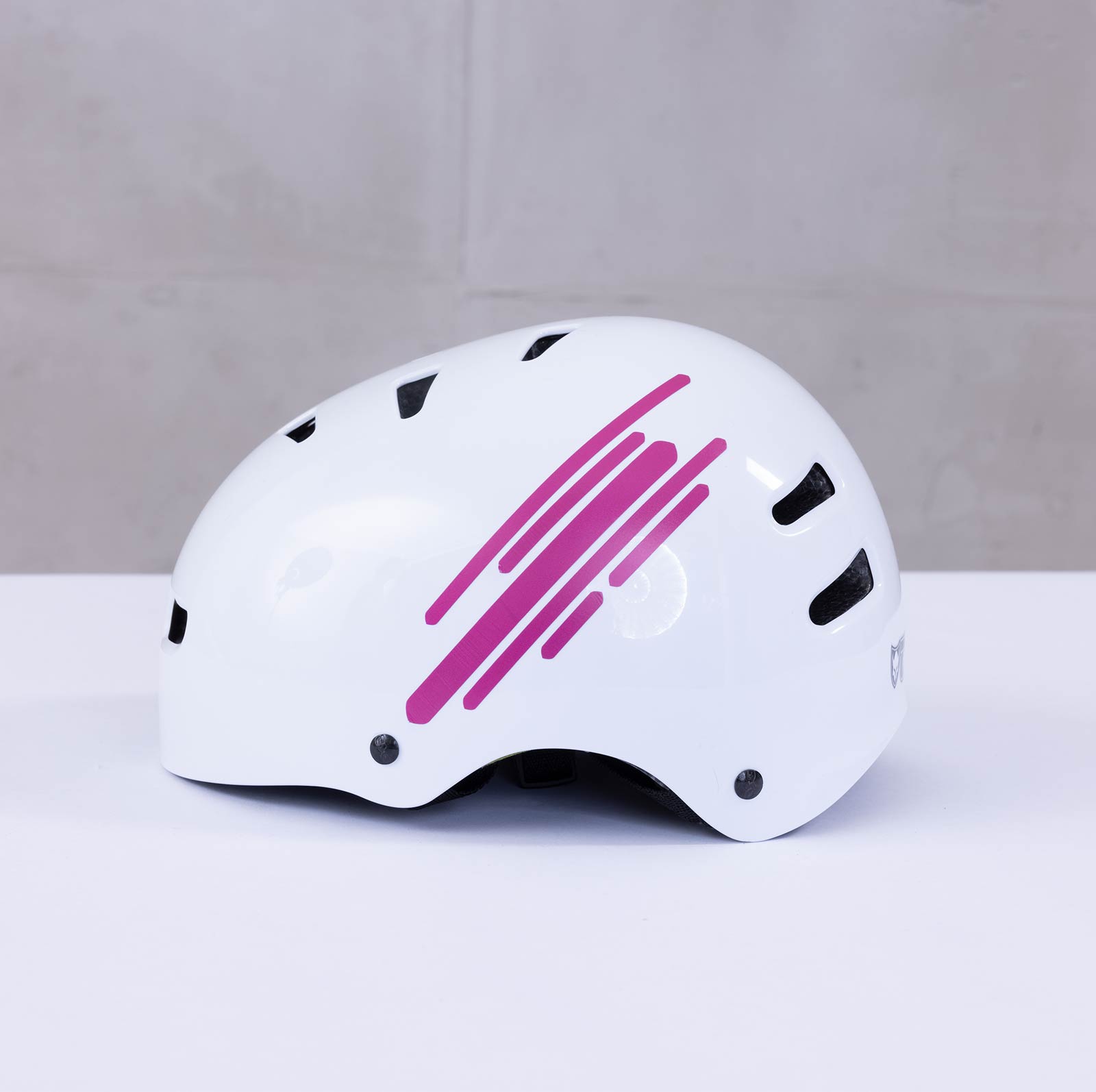 Ein weißer Helm mit schwarzen Belüftungslöchern und rosa diagonalen Streifen an der Seite, platziert auf einer weißen Fläche vor hellgrauem Hintergrund.